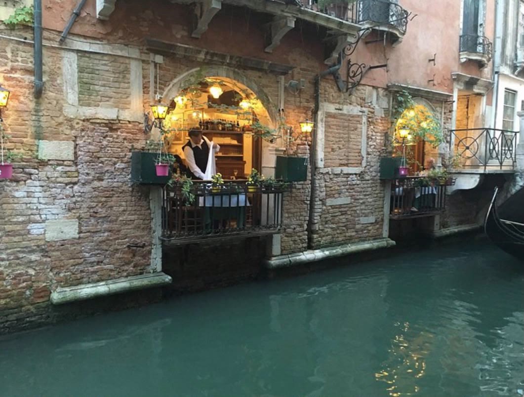 A candle-lit canal-side trattoria in Venice, geraniums spilling from the balcony, its warm interior mirrored in the green water below